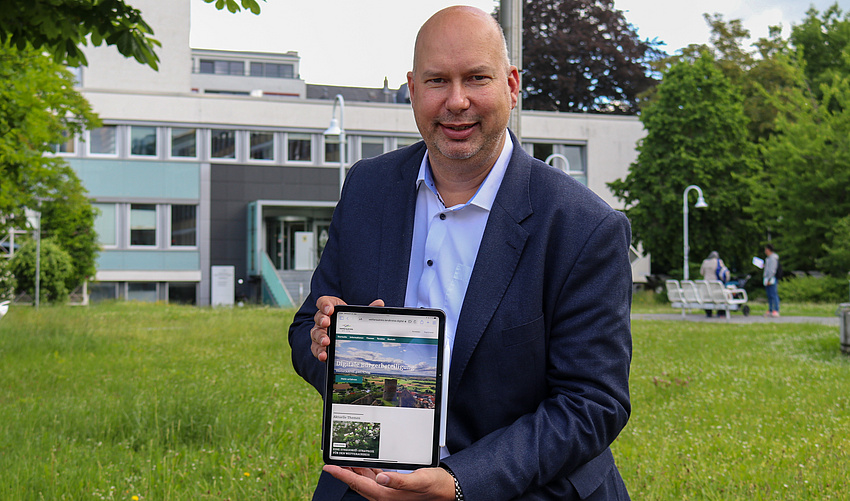 Ein Mann hält ein Tablet in die Kamera. Sie zeigt die Startseite der digitalen Bürgerbeteiligungsplattform. Im Hintergrund eine Wiese und ein Gebäude.