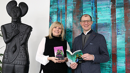 Ein Mann und eine Frau stehen nebeneinander und lächeln in die Kamera. Sie haben jeweils ein Buch in der Hand. Links neben ihnen eine lebensgroße schwarze Statue des Keltenfürsten. Im Hintrergrund ein buntes abstraktes Gemälde