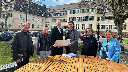 Sechs Menschen stehen an einem großen Holztisch, zwei Halten eine Akte in der Hand.