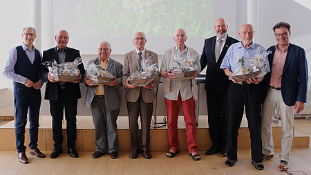 Sieben Männer stehen nebeneinander und lächeln in die Kamera. Fünf von ihnen haben einen kleinen Geschenkkorb in der Hand.