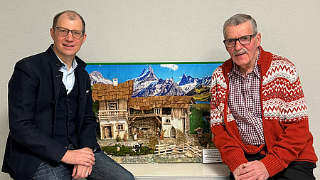 Zwei Männer sitzen vor einer handgefertigten Krippe vor einem alpenländischen Panorama.