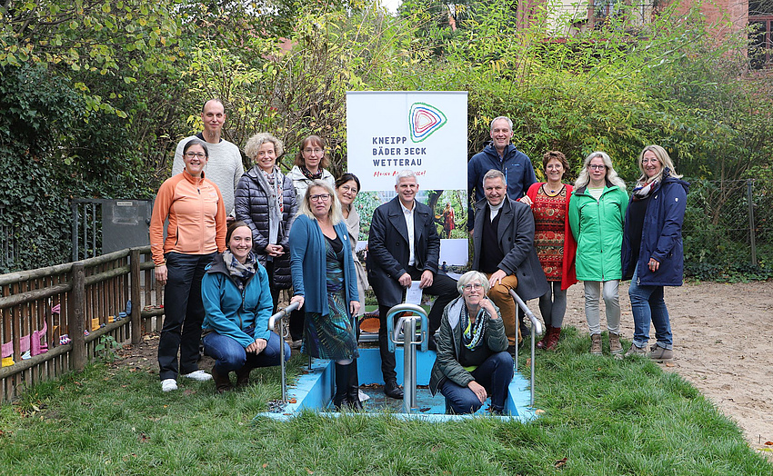 Eine Gruppe Männer und Frauen auf einer Wiese. Im Vordergrund eine Wassertretanlage. In ihr ist kein Wasser. Einige der Personen stehen in und am Rand des Beckens.