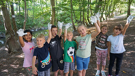 Jungen und Mädchen winken in die Kamera. Sie tragen jeweils an einer Hand weiße Handschuhe.