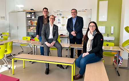 Blick in ein Klassenzimmer. Ein Mann und eine Frau sitzen jeweils auf einer Schulbank. Im Hintergrund stehen zwei Männer. Alle lächeln in die Kamera.