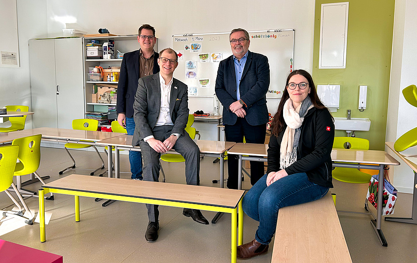 Blick in ein Klassenzimmer. Ein Mann und eine Frau sitzen jeweils auf einer Schulbank. Im Hintergrund stehen zwei Männer. Alle lächeln in die Kamera.