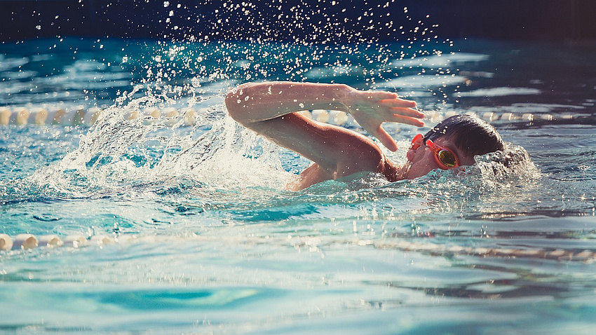 Ein Junge schwimmt im Wasser. Er krault undträgt eine Schwimmbrille.