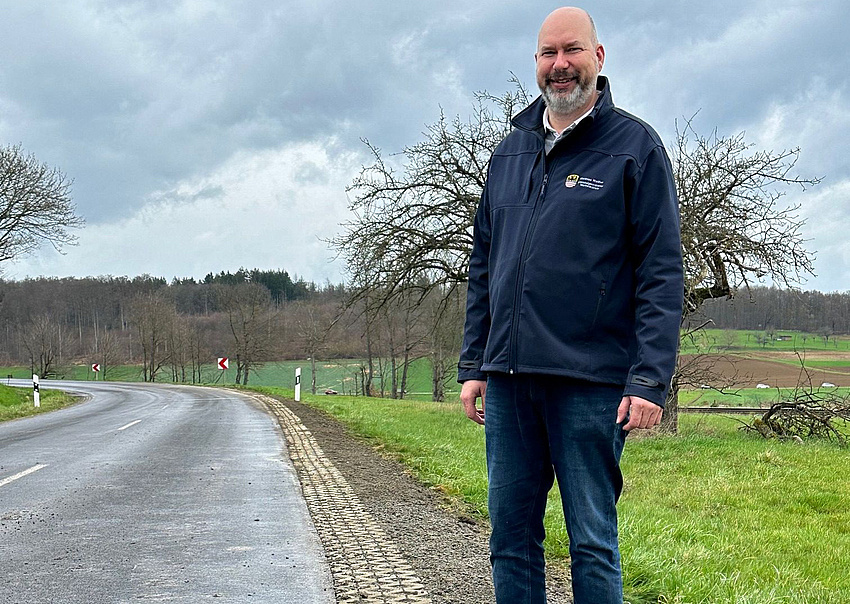 Ein Mann steht am Rand einer Straße, die durch die Natur führt. Am Straßenrand sind kleine Steine zu einem Gitter verlegt.