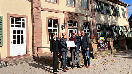 Zwei Männer halten ein Schriftstück in Händen. Neben ihnen ein Mann und eine Frau. Im Hintergrund ein Schlossgebäude