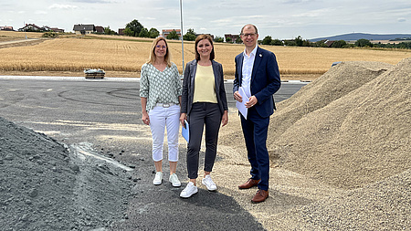 Ein Mann und zwei Frauen stehen auf einer Baustelle. Links und rechts sind Sandhaufen zu sehen, im Hintergrund ein Getreidefeld.