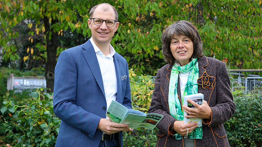 Ein Mann und eine Frau stehen vor einer grünen Hecke und halten einen Flyer in der Hand.