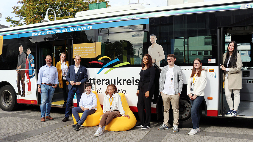 Mehrere Personen stehen vor einem Bus. Auf dem Bus sind diese Personen abgebildet. Sie werben für den Wetteraukreis als Arbeitgeber.