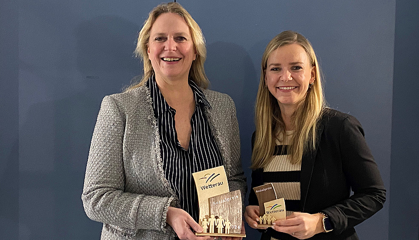 Zwei Frauen stehen nebeneinander. Sie halten ein kleines Kunstwerk aus Holz in der Hand. Es stellt den Wetterauer Sozialpreis dar.