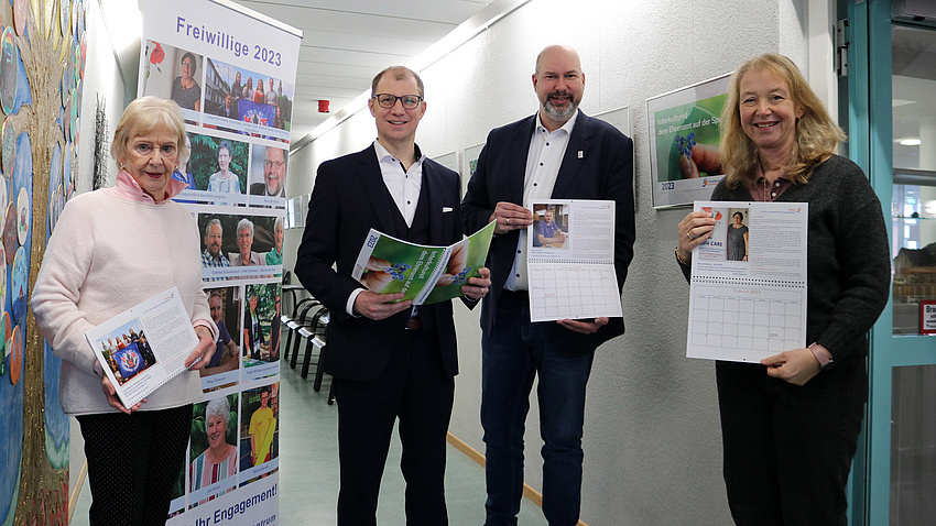 Zwei Frauen und zwei Männer haben jeweils einen Kalender in der Hand. Sie klächeln in die Kamera. Im Hintergrund ein großer Aufsteller mit einzelnen Bildern von Personen. Darüber die Überschrift: Ehrenamt 2023