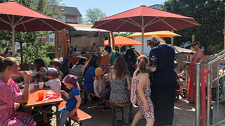 Erwachsene und Kinder frühstücken gemeinsam. Sie sitzen unter Sonnenschirmen an Bierzeltgarnituren.