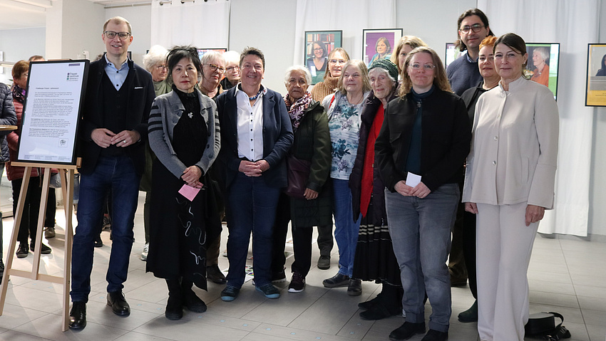 Zu sehen sind eine Gruppe von Frauen und zwei Männer. Sie lächeln in die Kamera. Im Hintergrund sind Fotografien von Frauen zu sehen.