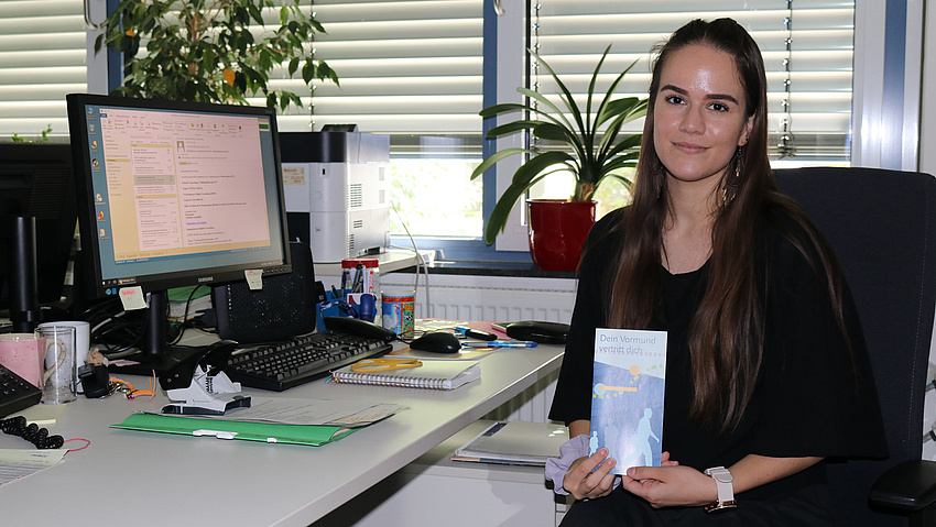 Eine junge Frau sitzt an ihrem Schreibtscih. Sie blickt ind die Kamera und hält ein Faltblatt in der Hand.