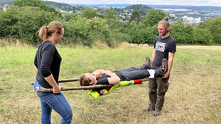 Ein Mann und eine Frau tragen eine selbstgebaute Trage aus dicken Ästen. Darauf liegt eine weitere Person, die so tut, als sie verletzt.