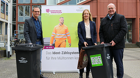 Drei Verantwortliche stehen mit zwei Mülltonnen vor einem großen Plakat des Abfallwirtschaftsbetriebes des Wetteraukreises mit der Aufschrift: Das Ident-Zählsystem für Ihre Mülltonnen kommt