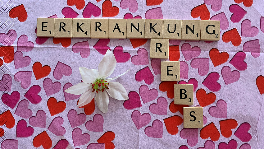 Die Wörter Erkrankung, Krebs mit Buchstaben  auf einem Untergrund mit rosa Herzen gelegt.