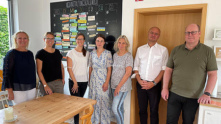 Zwei Männer und fünf Frauen stehen nebeneinander. Vor ihnen ein Küchentisch, im Hintergrund eine Wandtafel auf der Informationen stehen.