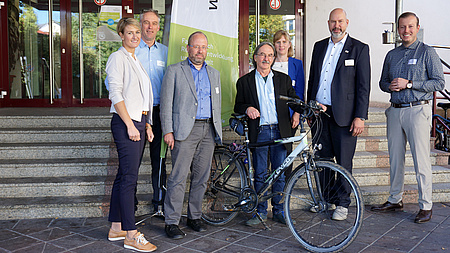 Eine kleine Personengruppe vor einer Treppe. Eine Person steht neben einem Fahrrad. Alle lächeln in die Kemare