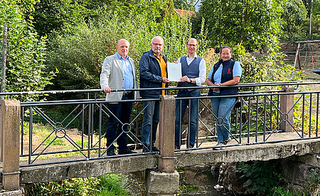 Drei Männer und eine Frau stehen auf einer Brücke. Im Hintergrund Bäume und Büsche