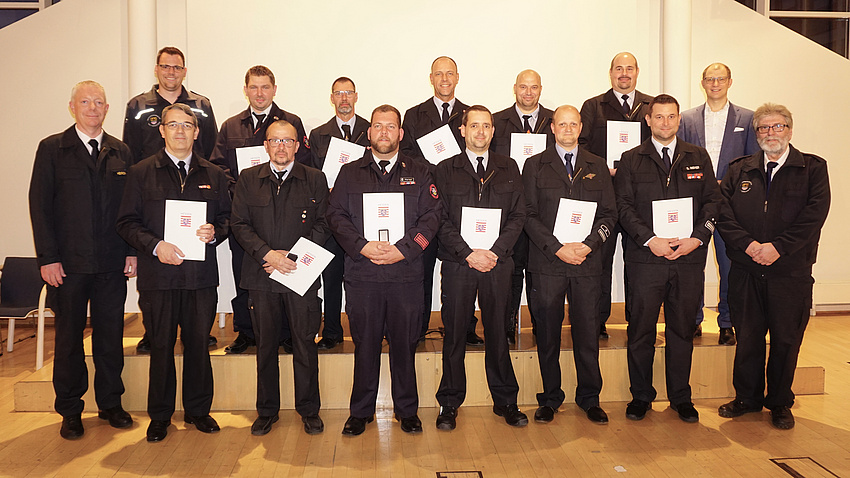 Eine größere Gruppe Männer steht in zwei Reihen hintereinander. Sie tragen Feuerwehruniformen, haben Urkunden in der Hand und blicken in die Kamera.