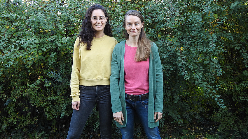 Zwei Frauen stehen im Freien vor einer grünen Hecke.