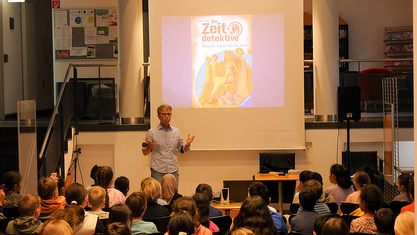 Kinder sitzen in Stuhlreihen in einer Bibliothek. Vor ihnen ein Mann, der zu ihnen spricht.