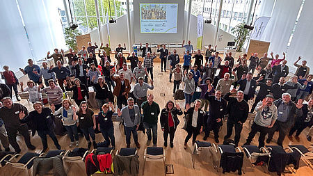 Eine große Menschengruppe steht in einem Saal. Alle haben die Hände gehoben und blicken nach oben zur Galerie, von der aus sie fotogtafiert werden.