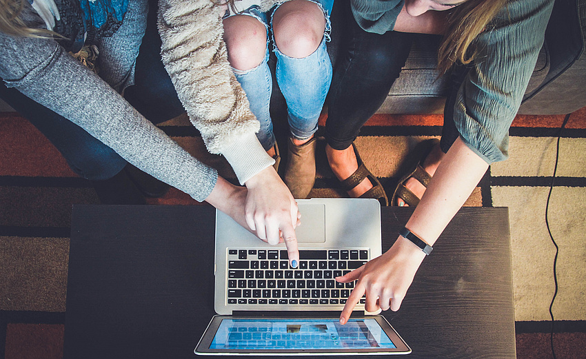 Drei junge Frauen sitzen auf einem Sofa, vor ihnen ein niederiger Tisch, auf dem ein Laptop steht. Eine Frau deutet auf den Bildschirm, die beiden anderen auf die Tastatur.