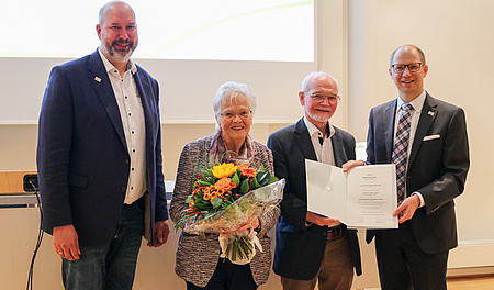 Drei Männer und eine Frau. Die Frau hält einen Blumenstrauß, zwei Männer halten eine Urkunde. Alle lächeln in die Kamera.