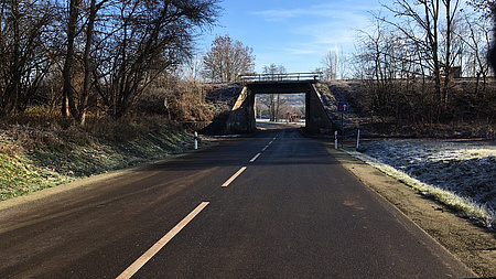 Eine asphaltierte Straße, links und rechts eine Wiese und Bäume. Im Vordergrund eine Brücke.