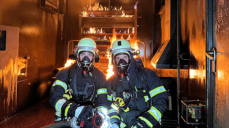 Zwei Feuerwehrleute in kompletter Ausrüstung knien in einem brennenden Container.