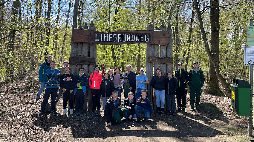 Eine Gruppe Jugendlicher im Wald. Sie stehen vor einem Tor aus Baumstämmen. Darüber ein Brett mit der Inschrift: Limesrundweg