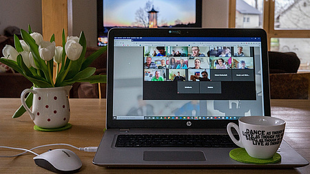 Blick auf einen aufgeklappten Laptop, eine Kaffeetasse, eine Vase mit Blumen.