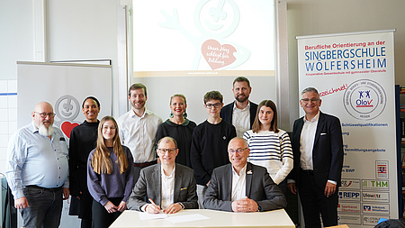 Mehrere Personen stehen nebeneinander. Vor ihnen sitzen zwei Männer an einem Tisch.