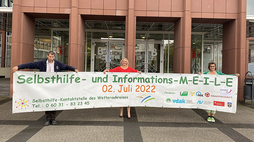 Von links: Dr. Reinhold Merbs, Erste Kreisbeigeordnete und Sozialdezernentin Stephanie Becker-Bösch und Anette Obleser. Alle halten ein 6 m langes Banner, welches auf die Selbsthilfe-MEILE hinweist.