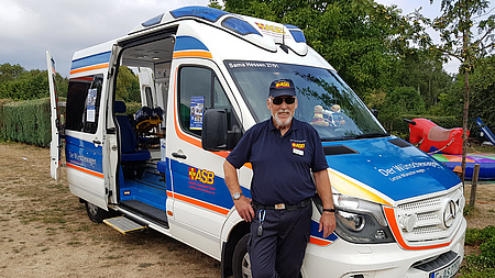 Ein Mann in dunkelblauer Dienstkleidung steht vor einem umgebauten Rettungswagen.