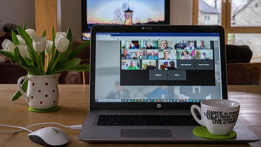 Ein aufgeklappter Laptop, auf dem Bildschirm sind die Gesichter aller Konferenzteilnehmer zu sehen. Daneben eine Tasse und eine Vase mit Tulpen.