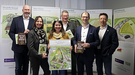 Zwei Frauen und vier Männer stehen nebeneinander. Zwei halten ein Buch in der Hand, zwei eine Broschüre und die Frau in der Mitte hält einen Bilderrahmen mit einem Plakat in die Kamera.