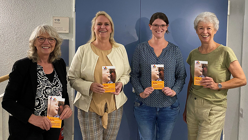 Vier Frauen stehen nebeinander und  lächeln  in die Kamera. Jede der Frauen hält ein Faltblatt in der Hand.