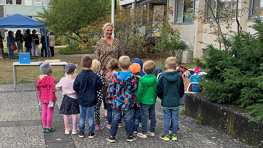 Eine Gruppe Kinder steht im Halbkreis mit dem Rücken zur Kamera vor einer Frau.e