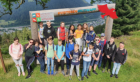 Eine Gruppe Jugendlicher steht auf einer Wiese. Im Hintergrund Bäume und Berge.