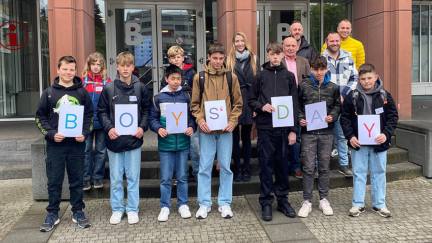 Zu sehen ist eine Gruppe von Jungen. Sie halten ausgedruckte Zettel mit Buchstaben hoch, die die Wörter "Boys' Day" ergeben.