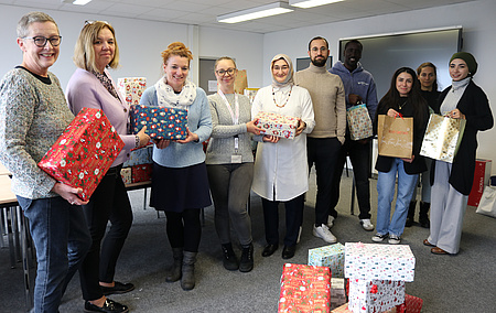 Mehrere Männer und Frauen stehen in einem Halbkreis nebeneinander. Sie halten weihnachtlich eingepackte Päckchen in den Händen.