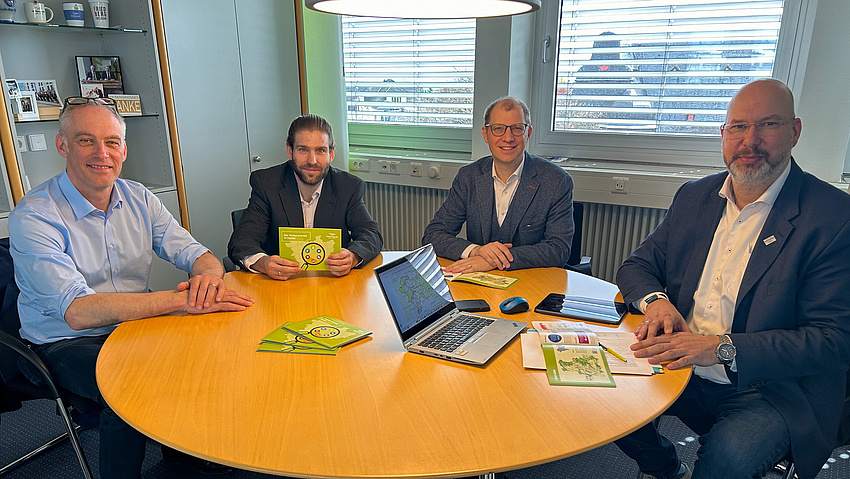 Vier Männer sitzen um einen runden Tisch. Auf dem Tisch liegen Broschüren und ein aufgeklappter Laptop.