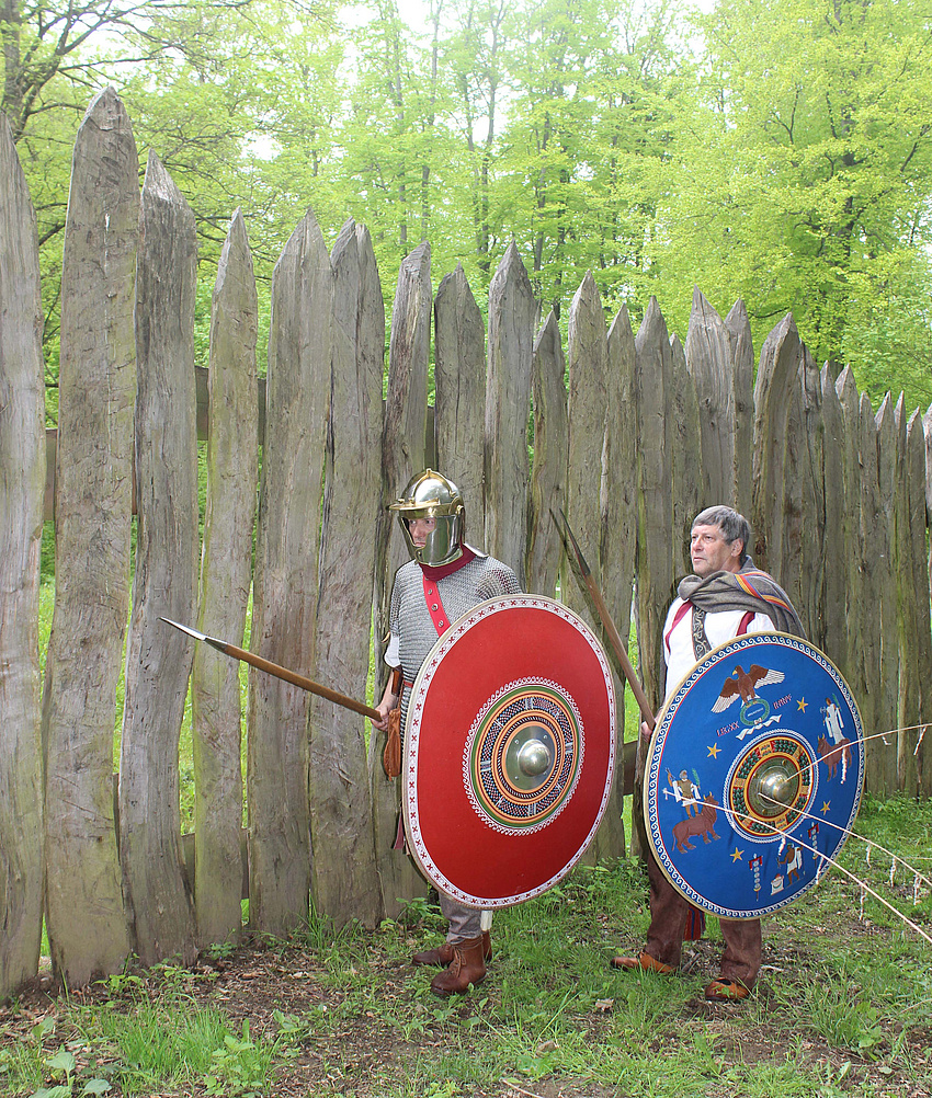 Zwei Personen in historischer römischer Gewandung, mit Schild und Bewaffnung. Neben ihnen ein Palisadenzaun.