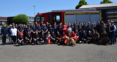 Eine größere Gruppe Männer und Frauen steht in einem Halbkreis. Vor ihnen weitere Personen in der Hocke. Im Vordergrund ein Hund. Im Hintergrund ein großes Fahrzeug der Feuerwehr.