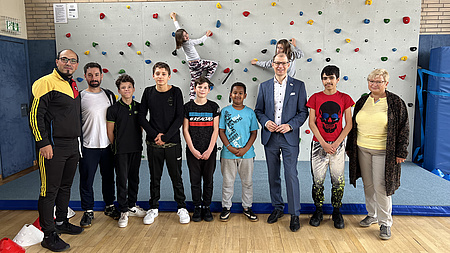 Kinder, Jugendliche und Erwachsene stehen nebeneinander in einer Turnhalle. Im Hintergrund eine Kletterwand, an der ein Mädchen klettert.